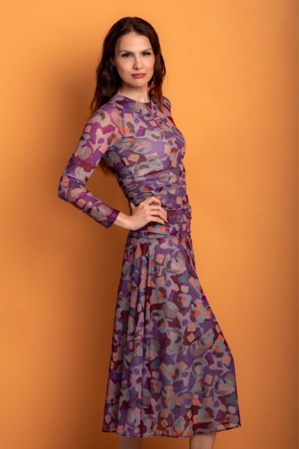 The woman wears a patterned midi dress with long sleeves and stands out against an orange background.