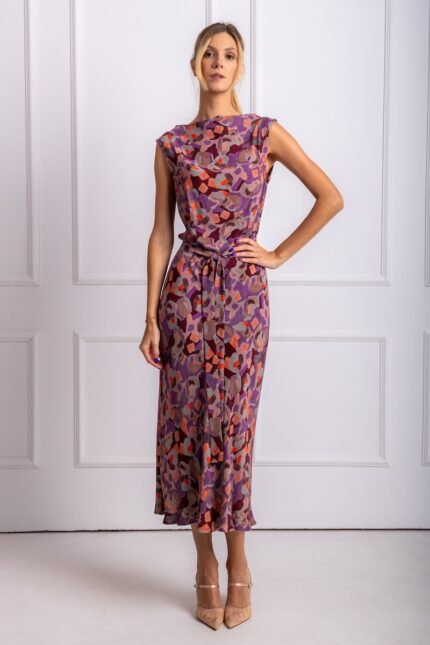 The blonde girl wears a patterned purple sleeveless midi dress. She is standing in front of a white wall.