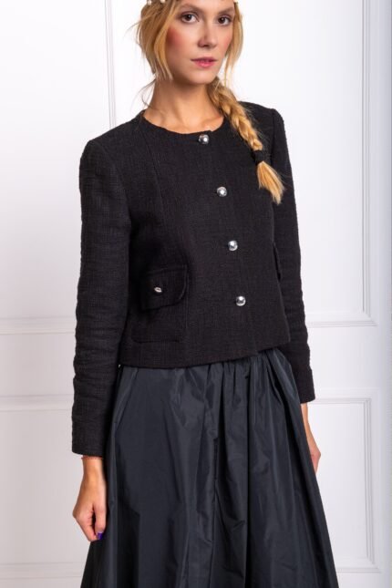 The blonde girl wears a black cotton blazer and a black midi skirt. She is standing in front of a white wall.