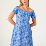 Plava letnja haljina sa belim floralnim printom, kratkih rukava i otvorenih ramena. Model nosi haljinu uz jednostavnu zlatnu ogrlicu