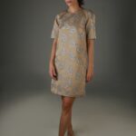 Žena nosi kratku bež haljinu sa kratkim rukavima i detaljima od plavog štrasa.