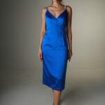 Žena nosi plavu satensku haljinu midi dužine sa bretelama i V izrezom.