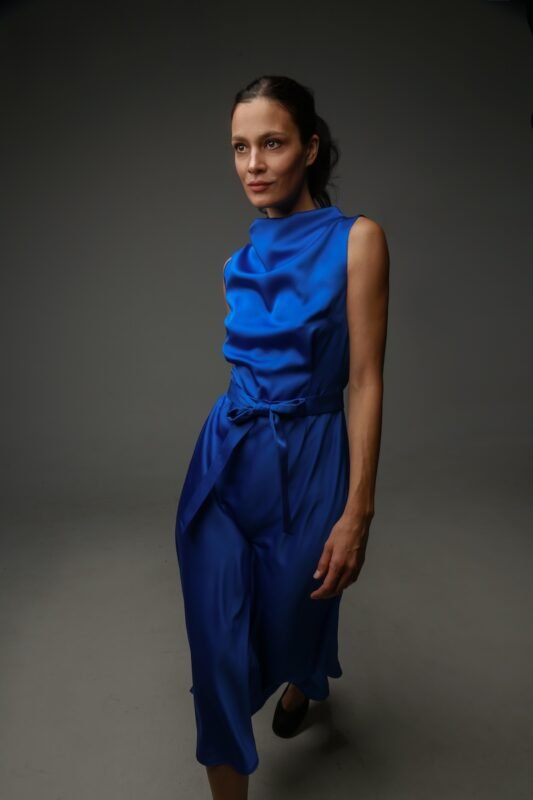 The woman wears a sleeveless midi length blue satin dress.