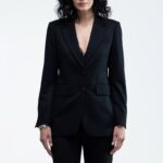 The woman wears a black waisted blazer and black skinny pants.