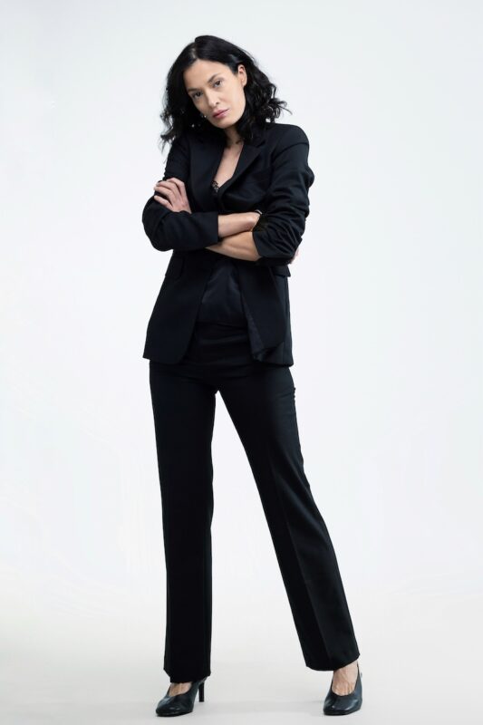 The woman wears a black waisted blazer and black skinny pants.