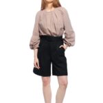 Devojka nosi duži crni šorts sa crnim pojasem oko struka u kombinaciji sa bež bluzom.